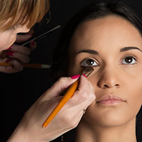 makeup Une maquilleuse applique minutieusement du fard à paupières sur les paupières d'une femme à l'aide d'un pinceau. La femme, les yeux ouverts et le regard légèrement levé, est assise sur un fond sombre. Ce moment pourrait facilement être immortalisé par un photographe de Genève.