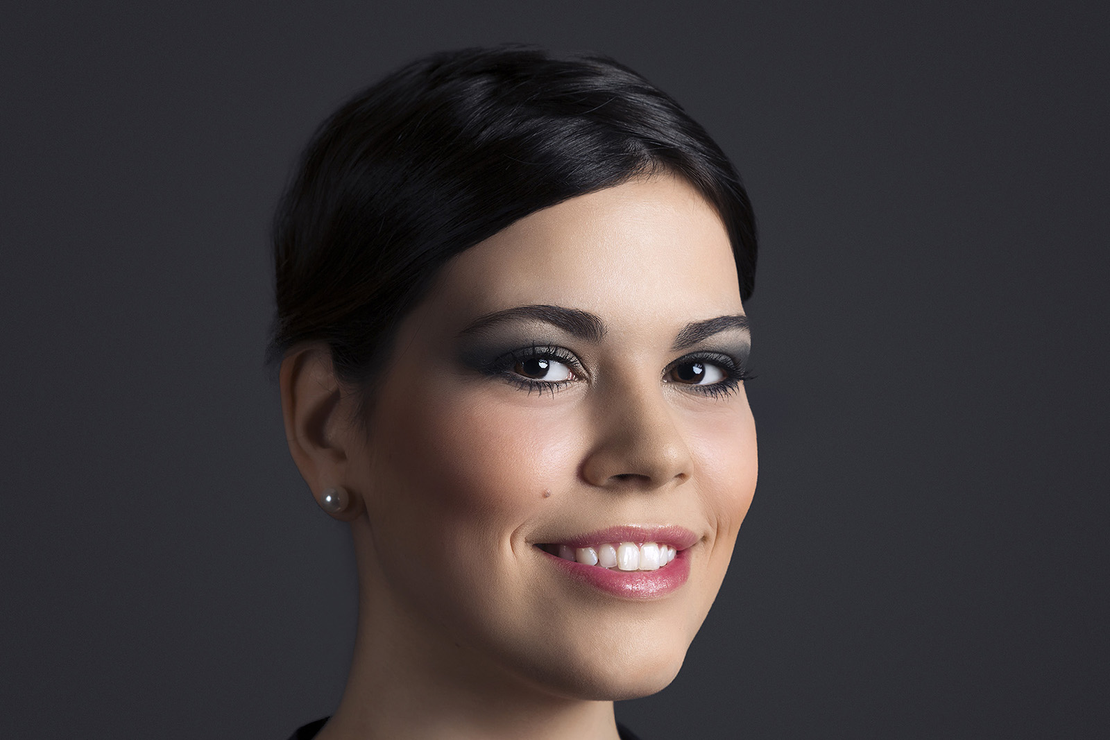 Une femme aux cheveux courts et foncés sourit doucement à l'objectif. Elle porte un maquillage foncé pour les yeux et un rouge à lèvres rose, avec des boucles d'oreilles en perles visibles. L'arrière-plan uni et sombre met en valeur son visage. Ce cliché captivant témoigne du talent du photographe Genève.