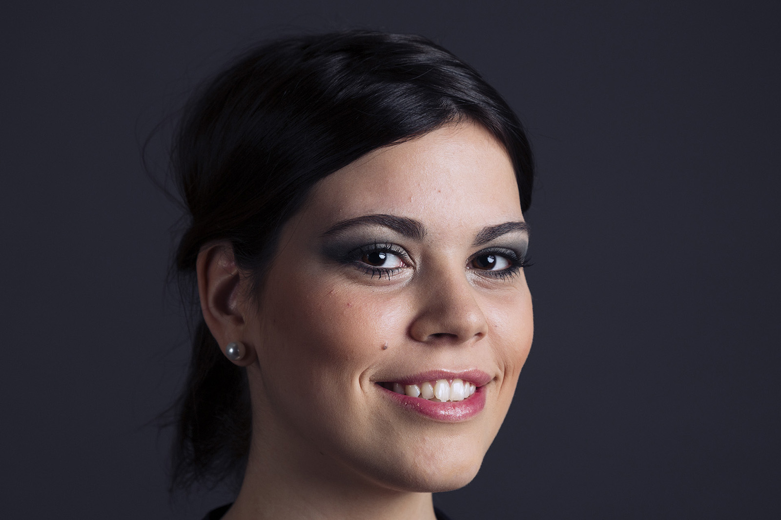 Une femme aux cheveux noirs sourit à l'objectif. Elle porte des boucles d'oreilles en perles et a un maquillage naturel avec un fard à paupières foncé. L'arrière-plan est sombre et uni, mettant en valeur son visage, parfaitement capturé par la photographe Geneve.