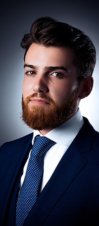 PRICELIST-BUSINESS Un jeune homme avec une barbe bien soignée et des cheveux noirs courts porte un costume bleu foncé, une chemise blanche et une cravate à pois bleus. Photographié par Photographe Geneve, il pose pour un portrait professionnel sur un fond sombre et dégradé.