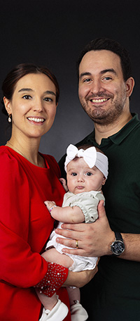 PRICELIST-FAMILY Un couple souriant tient dans ses bras un bébé vêtu de vêtements clairs et d'un bandeau blanc avec un noeud. La femme porte un haut rouge tandis que l'homme porte une chemise vert foncé. Ils se tiennent devant un fond sombre, magnifiquement capturé par le photographe Genève.