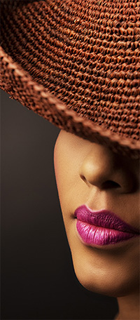 PRICELIST-FASHION Gros plan sur le visage d'une femme, partiellement recouvert d'un grand chapeau marron texturé. Ses lèvres, peintes d'un rouge à lèvres rose vif, sont bien visibles, créant un contraste saisissant avec sa peau éclatante et le fond sombre. Un superbe cliché capturé par le photographe Genève.