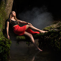 horsstudio Une femme en haut noir et jupe rouge est allongée sur un tronc d'arbre couvert de mousse, au-dessus d'un ruisseau brumeux et sombre. Captures par le photographe Genève, ses longs cheveux blonds tombent en cascade tandis que ses yeux restent fermés, donnant une apparence sereine et contemplative.