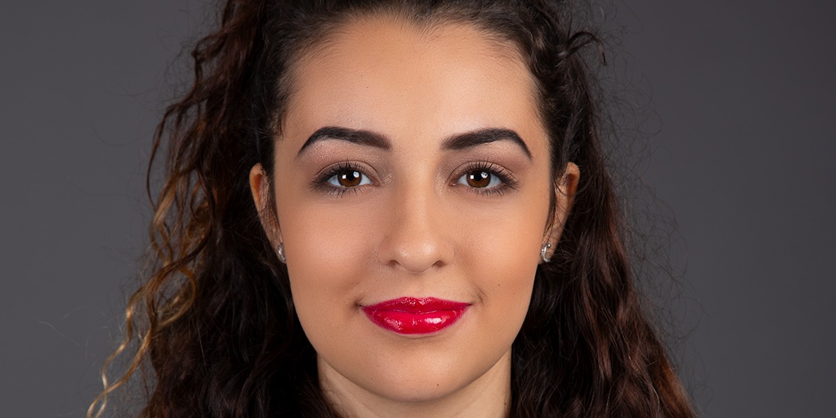 Une femme aux cheveux châtains longs et bouclés et au rouge à lèvres rouge vif fait face à l'objectif. Son expression est neutre et se détache sur un fond sombre et uni. Elle porte de petites boucles d'oreilles. Prise par le photographe Geneve, l'image respire la simplicité et l'élégance.
