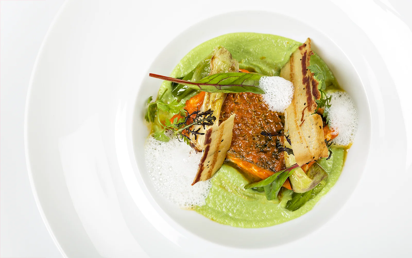 Un plat gastronomique composé d'un filet saisi sur un lit de purée verte, garni d'écume, de légumes grillés, de verdure fraîche et d'herbes, élégamment présenté sur un plat blanc.