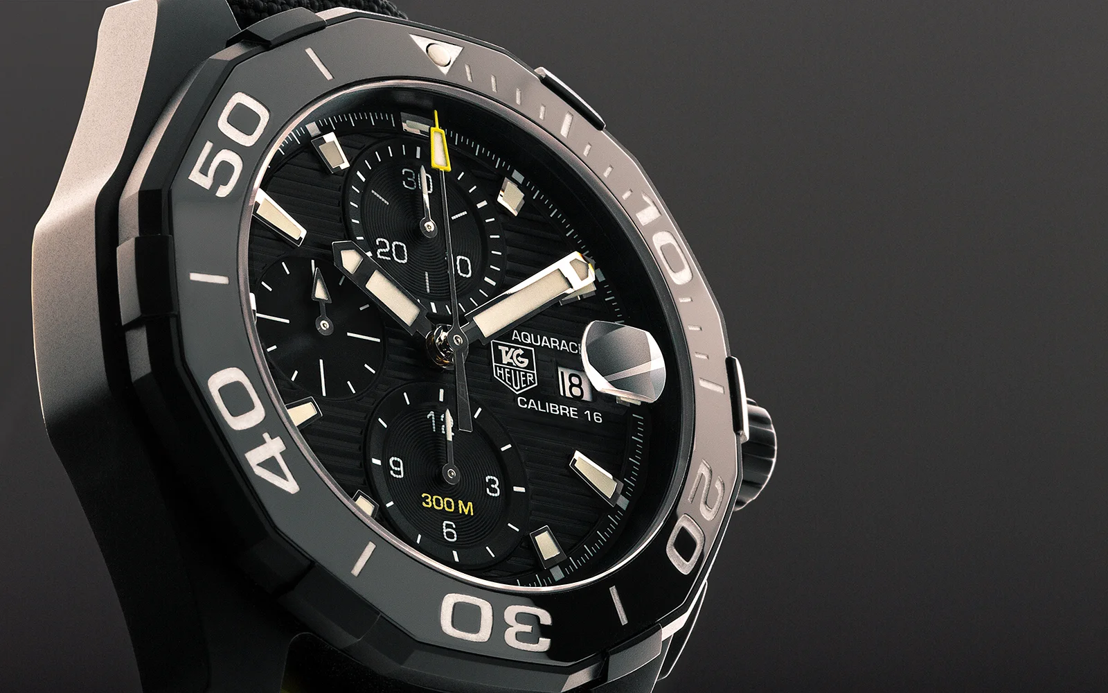 Gros plan sur une montre TAG Heuer Aquaracer Calibre 16 noire avec une lunette noire, des chiffres et des index blancs, trois cadrans auxiliaires, un guichet de date à 3 heures, et des accents jaunes sur la trotteuse et le marquage 300M.