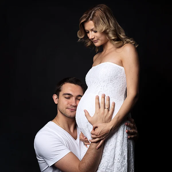 Un homme en chemise blanche s'agenouille, touche délicatement le ventre d'une femme enceinte et y appuie son visage. Capturée par Photographe Geneve, la femme vêtue d'une robe blanche en dentelle se tient debout et sourit doucement, tenant la main de l'homme sur un fond sombre.