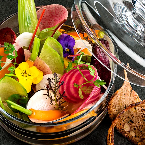 Un bol en verre rempli de légumes colorés coupés en fines tranches, de fleurs comestibles et d'herbes fraîches, avec un couvercle transparent partiellement ouvert. Des tranches de pain grillé reposent à côté du bol sur une surface sombre, comme l'a photographié le photographe Suisse Sylvain Rivarolo.