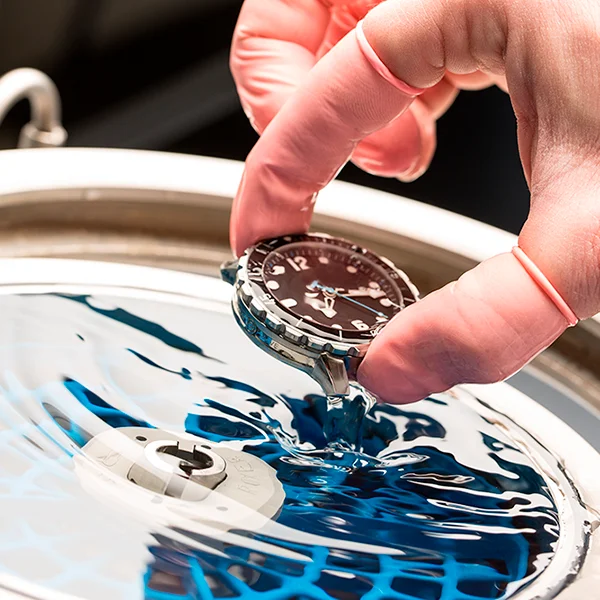 Une main gantée plonge une montre-bracelet dans un récipient d'eau peu profond, démontrant ainsi l'étanchéité de la montre. Des ondulations bleues se forment à la surface de l'eau, tandis que la scène est artistiquement capturée par le Photographe Geneve.